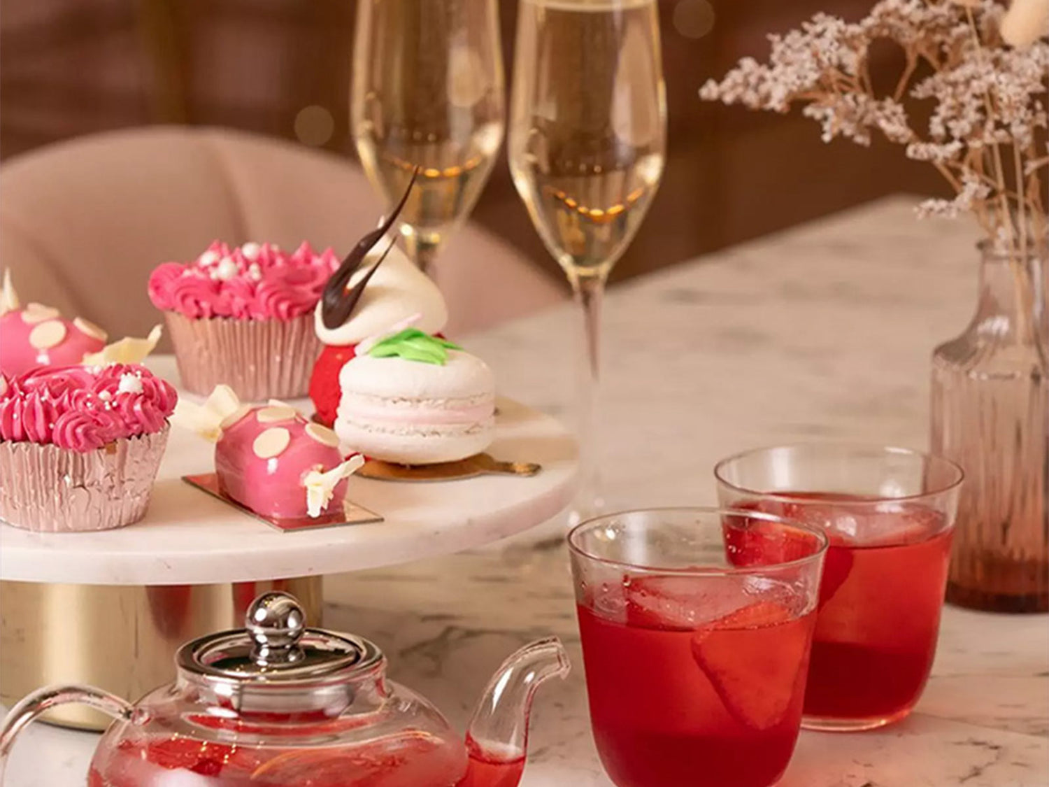 Afternoon tea setup with pink cupcakes, macarons, and cocktails on a marble table with champagne and flowers.