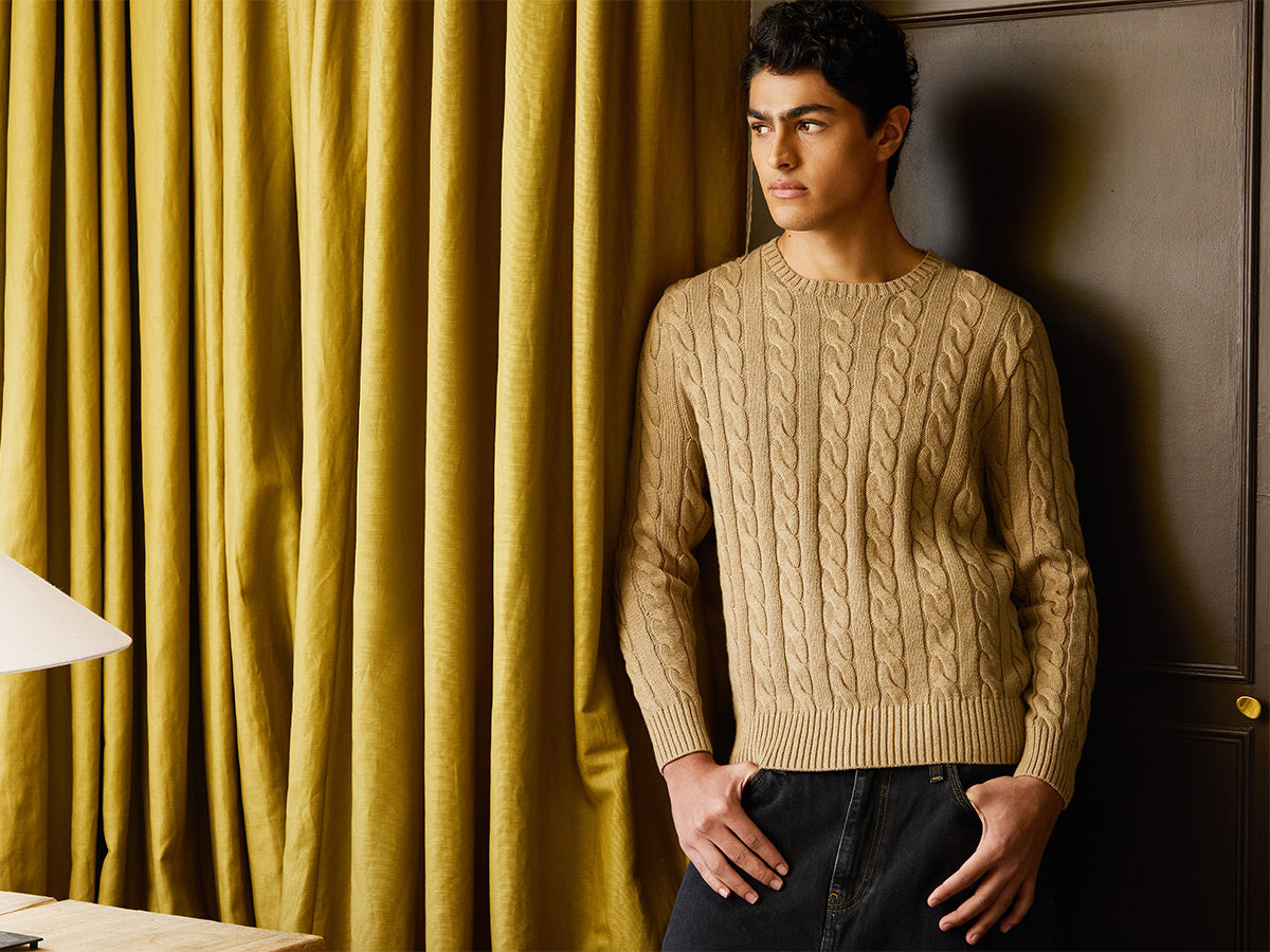 Man leaning against a yellow curtain wearing a beige knit jumper